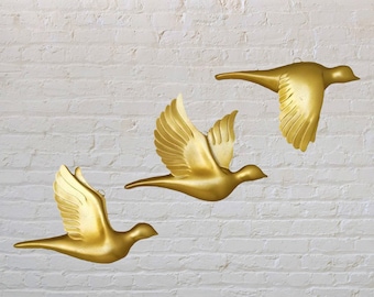 May include: Three gold-colored metal bird sculptures in flight, mounted on a white brick wall.
