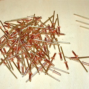 May include: A large pile of gold-colored metal rivets with a few rivets laying separately on a light brown surface.