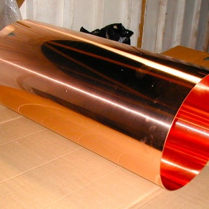 May include: A long, rolled sheet of copper metal with a shiny, reflective surface. The copper is a reddish-orange color.