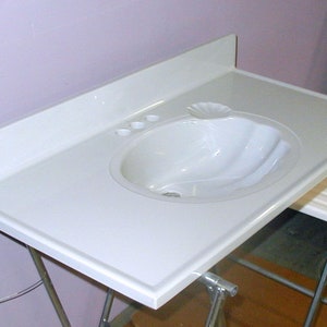 May include: White bathroom vanity countertop with a white porcelain sink and three holes for fixtures. The countertop is supported by a metal stand.