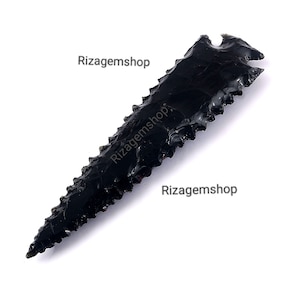 May include: A black obsidian arrowhead with a serrated edge. The arrowhead is approximately 5 inches long. Rizagemshop is printed on the image.