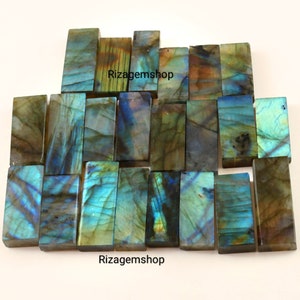 May include: A collection of 18 labradorite gemstone rectangular cabochons. The stones are a mix of blue, green, and gold colors with iridescent flashes. The stones are arranged in rows and are ready to be used in jewelry making.