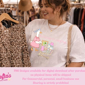 Peut inclure: Un t-shirt blanc avec un dessin d'oie poussant un chariot rempli d'articles. L'oie porte une robe rose et un nœud bleu. Un chemisier à imprimé léopard est suspendu à un cintre en bois au premier plan.