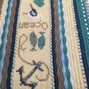 May include: A crocheted throw blanket with a beige background and blue, brown, and green stripes. The centre of the blanket features a cross-stitch design with anchors, fish, seagulls, and the words "We Do Beach".