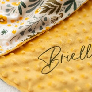 May include: A yellow baby blanket with a textured surface and the embroidered name "Brielle" in a cursive font. The blanket features a floral pattern in cream, yellow, and green on the reverse side, creating a soft and cozy appearance.