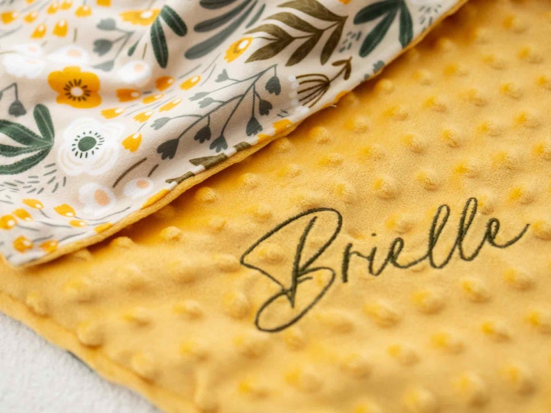 May include: A yellow baby blanket with a textured surface and the embroidered name "Brielle" in a cursive font. The blanket features a floral pattern in cream, yellow, and green on the reverse side, creating a soft and cozy appearance.