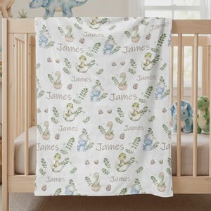 Könnte beinhalten: Weiße Babydecke mit einem sich wiederholenden Muster aus Aquarell-Dinosauriern, Eiern und Blättern. Der Name "James" ist in einer Schreibschrift gedruckt. Die Decke ist über ein Holzbett drapiert.