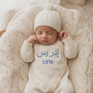 May include: A cream-colored knitted long-sleeve romper with a side button closure and a matching pom-pom beanie. The romper features blue embroidered text in both Arabic script and the English name Idris centered on the chest area.