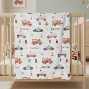 May include: A white baby blanket featuring a pattern of red fire trucks, ambulances, and police cars. The blanket also includes illustrations of traffic cones, first aid kits, and the name "Jackson" in brown script. The blanket is draped over a crib.