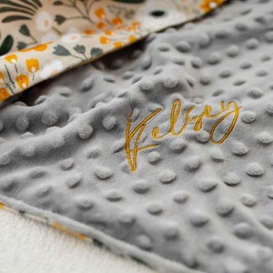 May include: A grey, dimpled baby blanket embroidered with the name "Kelsey" in gold thread. The blanket features a floral patterned border with yellow and white flowers and green leaves.