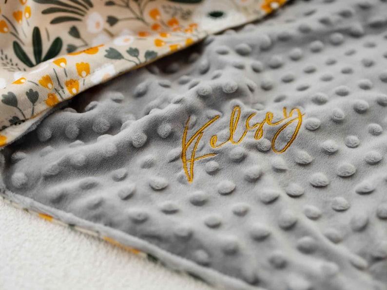 May include: A grey, dimpled baby blanket embroidered with the name "Kelsey" in gold thread. The blanket features a floral patterned border with yellow and white flowers and green leaves.