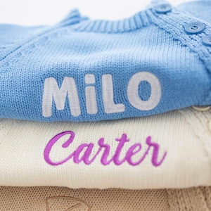 May include: Close-up of three folded knit cardigans. The top cardigan is light blue with the name "Milo" embroidered in white. The middle cardigan is cream-colored with the name "Carter" embroidered in purple. The bottom cardigan is tan.