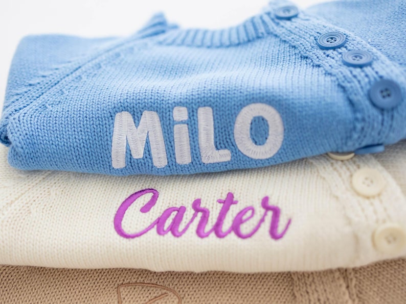 May include: Close-up of three folded knit cardigans. The top cardigan is light blue with the name "Milo" embroidered in white. The middle cardigan is cream-colored with the name "Carter" embroidered in purple. The bottom cardigan is tan.