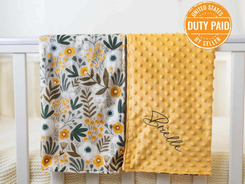 May include: Two baby blankets on a white cot. One has a floral pattern with yellow, white, and green. The other is a textured, yellow blanket with the name "Brielle" in script. The blankets appear soft and cosy, suitable for a baby.