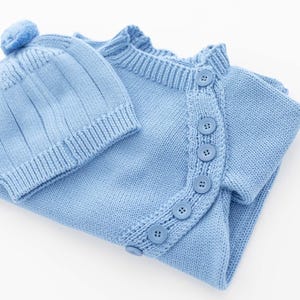 May include: A light blue knitted baby outfit, including a button-up cardigan and a matching beanie with a pom-pom. The set is made of soft knit fabric and features a classic design, perfect for newborns.