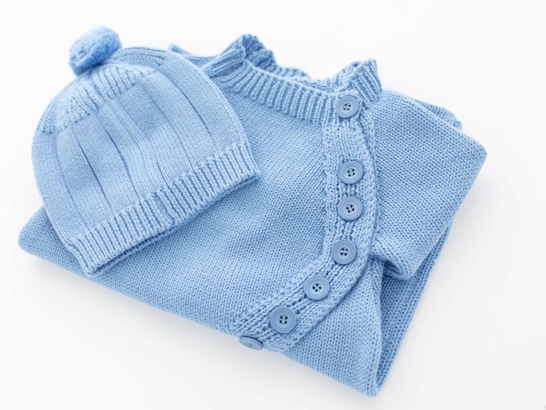 May include: A light blue knitted baby outfit, including a button-up cardigan and a matching beanie with a pom-pom. The set is made of soft knit fabric and features a classic design, perfect for newborns.