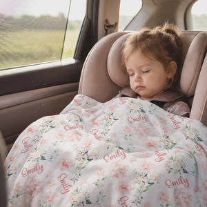 May include: A sleeping child in a car seat is covered with a white floral blanket. The blanket has a repeating pattern of pink flowers and the name "Emily" in a cursive font. The car seat is a light pink color.