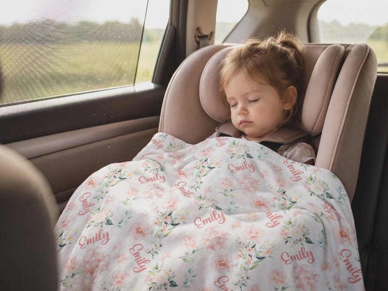 May include: A sleeping child in a car seat is covered with a white floral blanket. The blanket has a repeating pattern of pink flowers and the name "Emily" in a cursive font. The car seat is a light pink color.