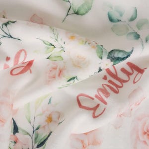 May include: A white baby blanket with a floral design featuring pink roses and green leaves. The name "Emily" is written in a cursive font in pink. The blanket is soft and plush, with a delicate, watercolor-like aesthetic.