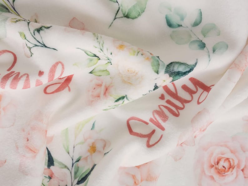 May include: A white baby blanket with a floral design featuring pink roses and green leaves. The name "Emily" is written in a cursive font in pink. The blanket is soft and plush, with a delicate, watercolor-like aesthetic.