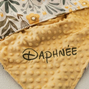 May include: A yellow baby blanket with a textured, dotted surface. The blanket has the name "DAPHN&Eacute;E" embroidered in dark green thread. The top layer features a floral pattern with yellow and green flowers and leaves.
