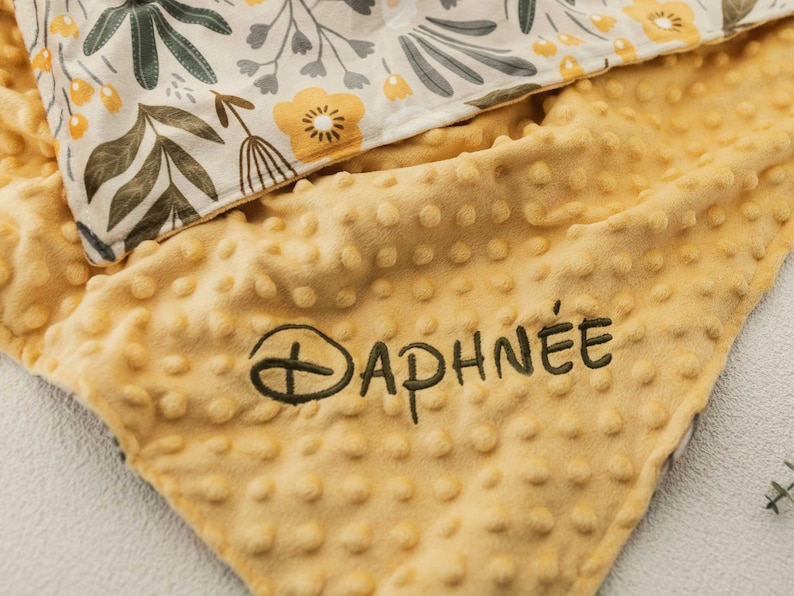 May include: A yellow baby blanket with a textured, dotted surface. The blanket has the name "DAPHN&Eacute;E" embroidered in dark green thread. The top layer features a floral pattern with yellow and green flowers and leaves.