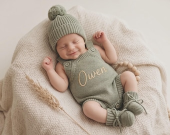May include: A sage green knitted baby romper featuring the name Owen embroidered in cream thread on the chest, paired with a matching ribbed beanie with a pom-pom and a pair of soft knitted booties with tie closures at the ankles.