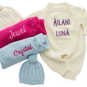 May include: An assortment of baby clothing items, including a cream-colored romper with the name "Ailani Luna" embroidered in purple. There are also pink and blue sweaters with the names "Jewel" and "Crystal" embroidered, along with matching hats.