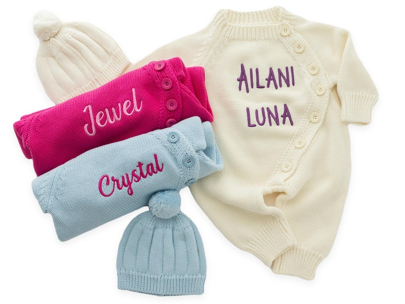 May include: An assortment of baby clothing items, including a cream-colored romper with the name "Ailani Luna" embroidered in purple. There are also pink and blue sweaters with the names "Jewel" and "Crystal" embroidered, along with matching hats.