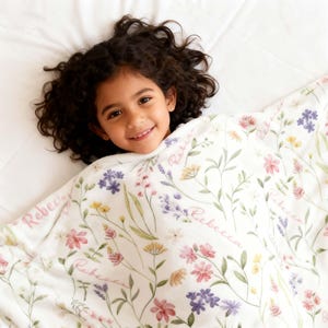 Op de afbeelding: Een kind lacht, gehuld in een witte deken met een bloemenpatroon. De deken heeft roze, paarse, gele en groene bloemen. De naam "Rebecca" is in roze script op de deken gedrukt.