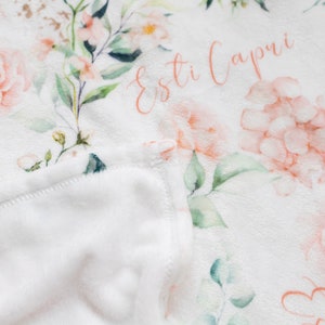 May include: A soft, white blanket with a floral pattern featuring pink roses and green leaves. The blanket has the name "Esti Capri" written in cursive. The edge of the blanket is folded over, revealing a plush, white interior.