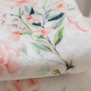 May include: A soft, white blanket with a floral pattern. The design features large, light pink roses and smaller peach-colored flowers, with green leaves and stems. The blanket has a plush texture, perfect for a baby.