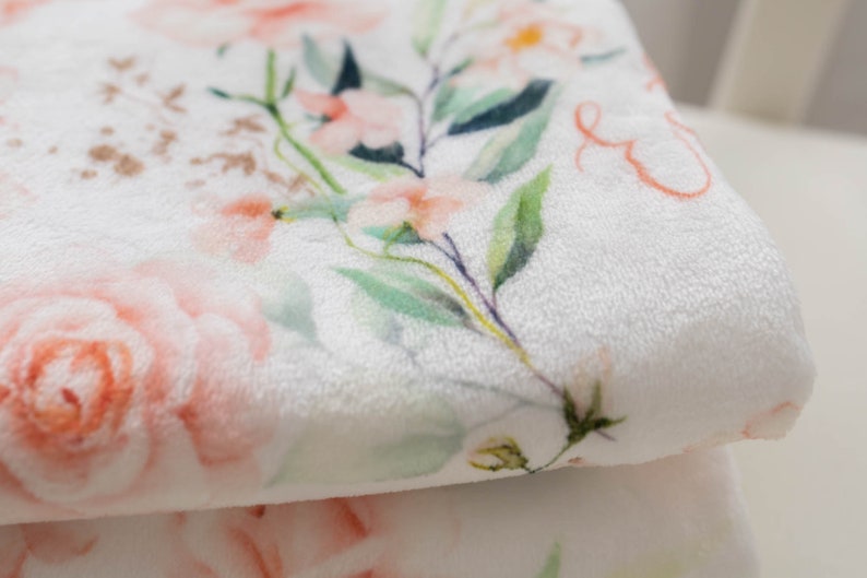 May include: A soft, white blanket with a floral pattern. The design features large, light pink roses and smaller peach-colored flowers, with green leaves and stems. The blanket has a plush texture, perfect for a baby.