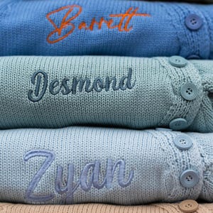 May include: Close-up of three personalized knit sweaters in shades of blue. Each sweater features an embroidered name in a contrasting color. The top sweater is light blue with an orange name, the middle is teal with a blue name, and the bottom is light blue with a light blue name.