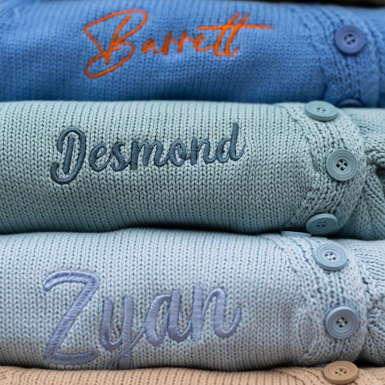 May include: Close-up of three personalized knit sweaters in shades of blue. Each sweater features an embroidered name in a contrasting color. The top sweater is light blue with an orange name, the middle is teal with a blue name, and the bottom is light blue with a light blue name.