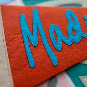 Puede incluir: Bandera de fieltro naranja con el nombre "Madie" en escritura turquesa. La bandera tiene un borde beige y est&aacute; cosida con hilo a juego. El fieltro tiene un aspecto texturizado.