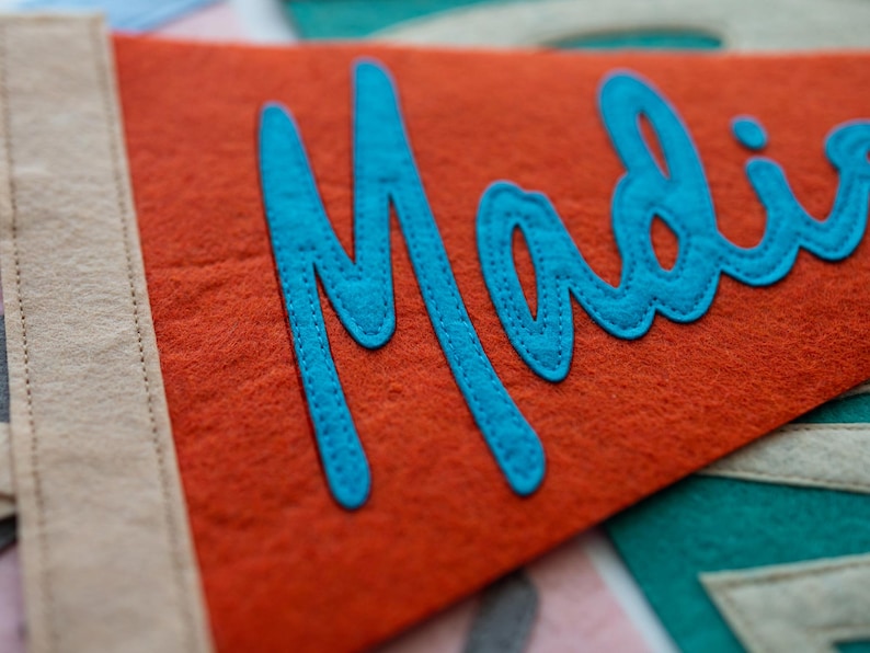 Puede incluir: Bandera de fieltro naranja con el nombre "Madie" en escritura turquesa. La bandera tiene un borde beige y est&aacute; cosida con hilo a juego. El fieltro tiene un aspecto texturizado.