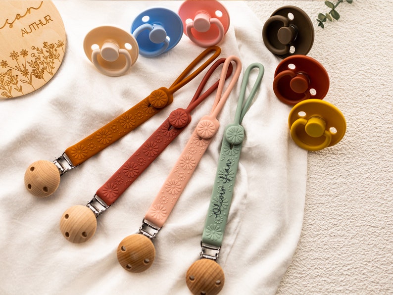 May include: Four silicone pacifier clips with wooden beads and floral patterns. The clips are in shades of brown, pink, green, and orange. The clips are attached to pacifiers with the names "Author" and "Dylan Jean" written on them.