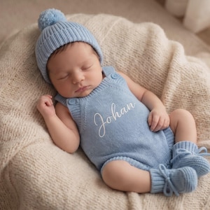 Op de afbeelding: Een lichtblauwe gebreide babyromper met de naam Johan in wit cursief schrift op de borst, gecombineerd met een bijpassende lichtblauwe muts met een pompon en een paar lichtblauwe gebreide babyslofjes met vetersluiting.