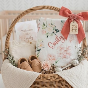 May include: A wicker basket filled with baby gifts, including a floral blanket with the name "Emily", a cream-colored sweater, and tan leather baby shoes. A pink bow and a wooden tag that says "Gift for Emily" adorn the basket.