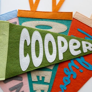 Puede incluir: Primer plano de varios banderines de fieltro en varios colores, incluyendo verde, naranja y rosa. El bander&iacute;n verde muestra el nombre "COOPER" en blanco, mientras que otros presentan nombres en diferentes fuentes y colores. Cada bander&iacute;n tiene un borde contrastante y una cinta para colgar.