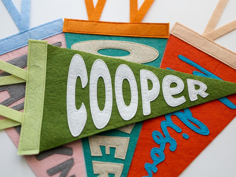 Puede incluir: Primer plano de varios banderines de fieltro en varios colores, incluyendo verde, naranja y rosa. El bander&iacute;n verde muestra el nombre "COOPER" en blanco, mientras que otros presentan nombres en diferentes fuentes y colores. Cada bander&iacute;n tiene un borde contrastante y una cinta para colgar.
