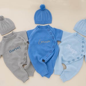 May include: Three baby outfits with matching hats. The outfits are in light blue, blue, and gray, each with a name embroidered on the front: "Harvey", "Lorenzo", and "Thiago". The hats have a ribbed design and a small pom-pom on top.
