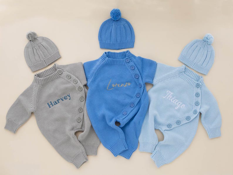 May include: Three baby outfits with matching hats. The outfits are in light blue, blue, and gray, each with a name embroidered on the front: "Harvey", "Lorenzo", and "Thiago". The hats have a ribbed design and a small pom-pom on top.