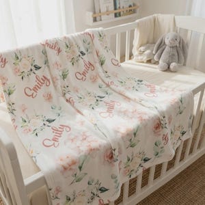 May include: A white baby crib with a floral-patterned blanket draped over the side. The blanket features pink roses, green leaves, and the name "Emily" in a cursive font. A gray stuffed bunny sits inside the crib.