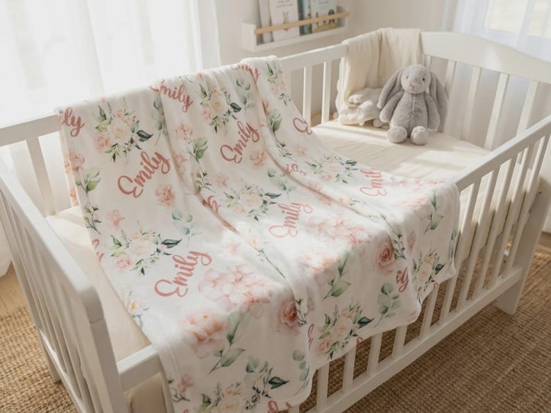 May include: A white baby crib with a floral-patterned blanket draped over the side. The blanket features pink roses, green leaves, and the name "Emily" in a cursive font. A gray stuffed bunny sits inside the crib.
