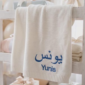 May include: A cream-colored knitted baby blanket featuring a ribbed texture and a ribbed border. The blanket is embroidered with the name Yunis in blue thread, written in both Arabic script and English text, centered on the lower portion of the fabric.