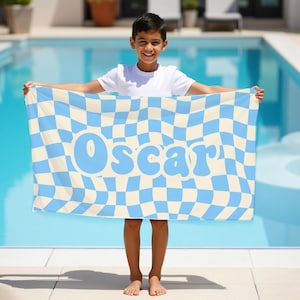May include: A beach towel with a blue and cream checkered pattern and the name "Oscar" in light blue. The towel is rectangular and held up by a person in front of a pool.