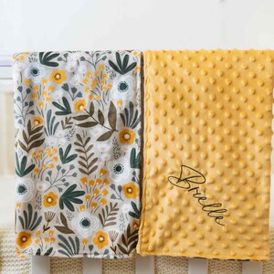 May include: Two baby blankets. One has a floral pattern with yellow, white, and green flowers on a beige background. The other is a textured, golden-yellow blanket with the name "Brielle" embroidered in black script.