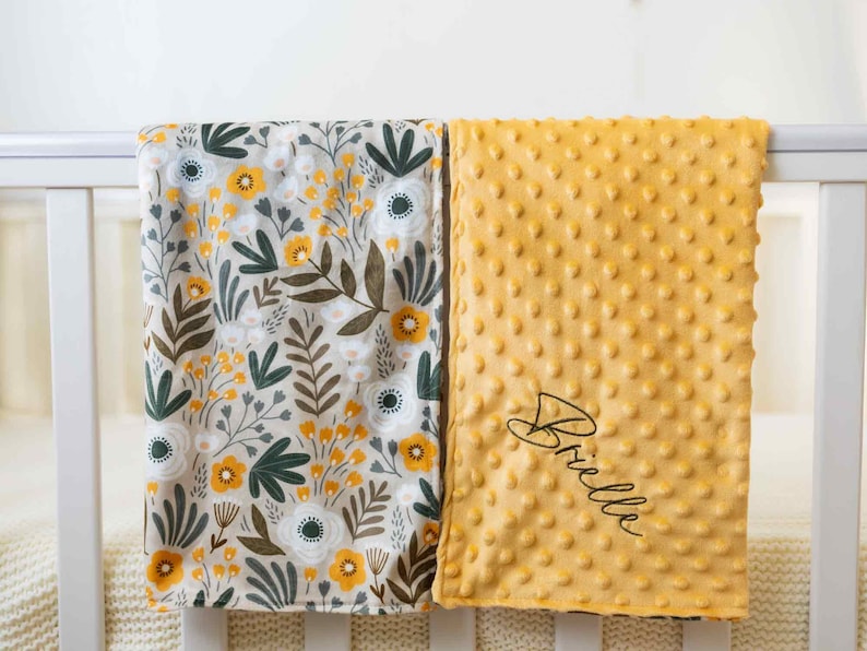 May include: Two baby blankets. One has a floral pattern with yellow, white, and green flowers on a beige background. The other is a textured, golden-yellow blanket with the name "Brielle" embroidered in black script.